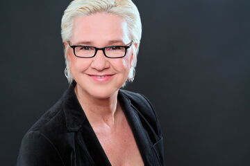 A person with short, blonde hair and glasses smiles warmly against a dark background, wearing a black blazer.