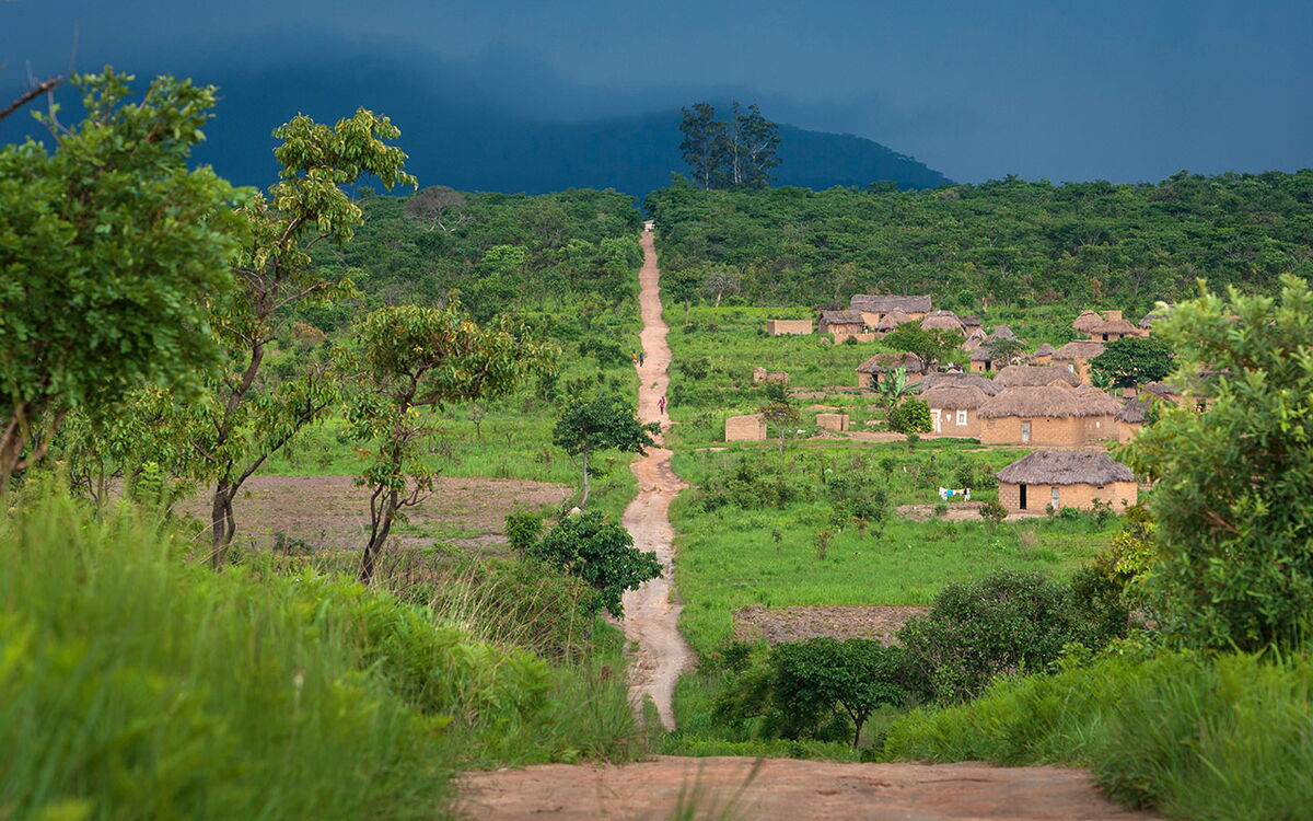 Ein schmaler Pfad führt durch eine üppige grüne Landschaft zu einem Dorf mit strohgedeckten Hütten unter dunklem Himmel.