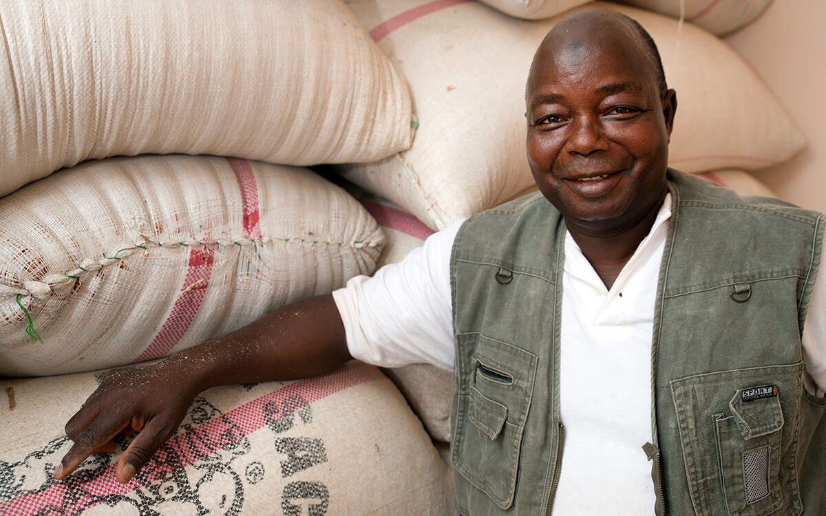 A smiling man in a vest leans against large sacks of goods, showcasing his work in Benin.