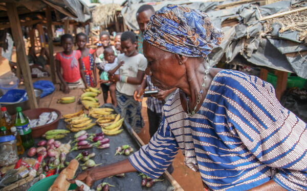 Eine ältere Frau in traditioneller Kleidung kauft Früchte auf einem lebhaften Marktstand, umgeben von anderen Marktbesuchern.