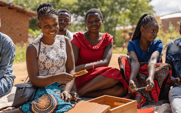 A group of smiling young people sit outdoors, engaging in a lively activity with a wooden box.