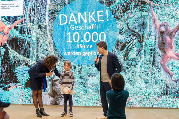 A woman and a man celebrate with a child in front of a backdrop announcing the success of planting 10,000 trees.