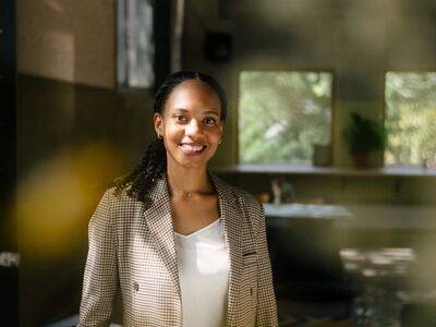 Eine lächelnde Frau im karierten Blazer steht in einem hellen Raum mit großen Fenstern.