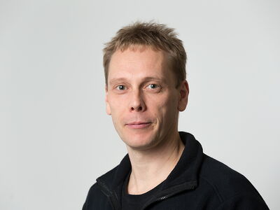 A person with short hair is looking at the camera against a plain background, wearing a dark jacket.