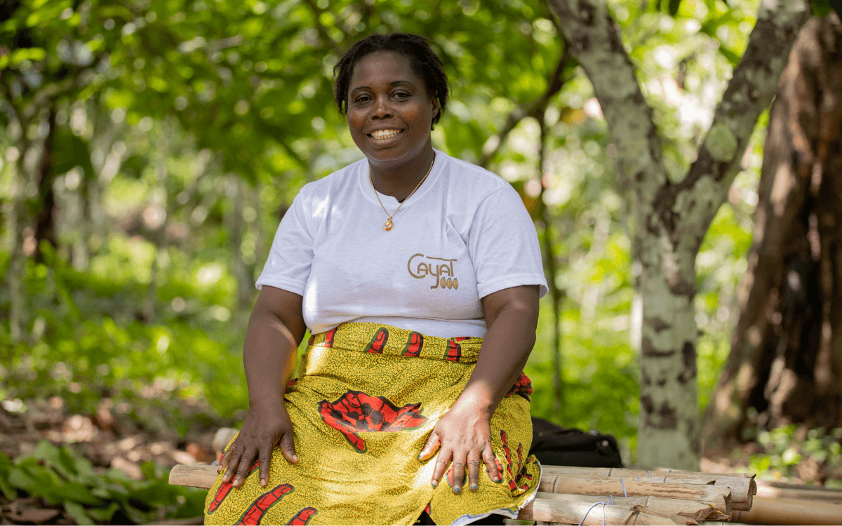 Eine Frau in traditioneller Kleidung sitzt lächelnd im Freien unter Bäumen in der Elfenbeinküste.