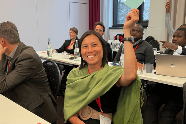 A smiling woman in a green shawl raises her hand enthusiastically during a conference meeting.