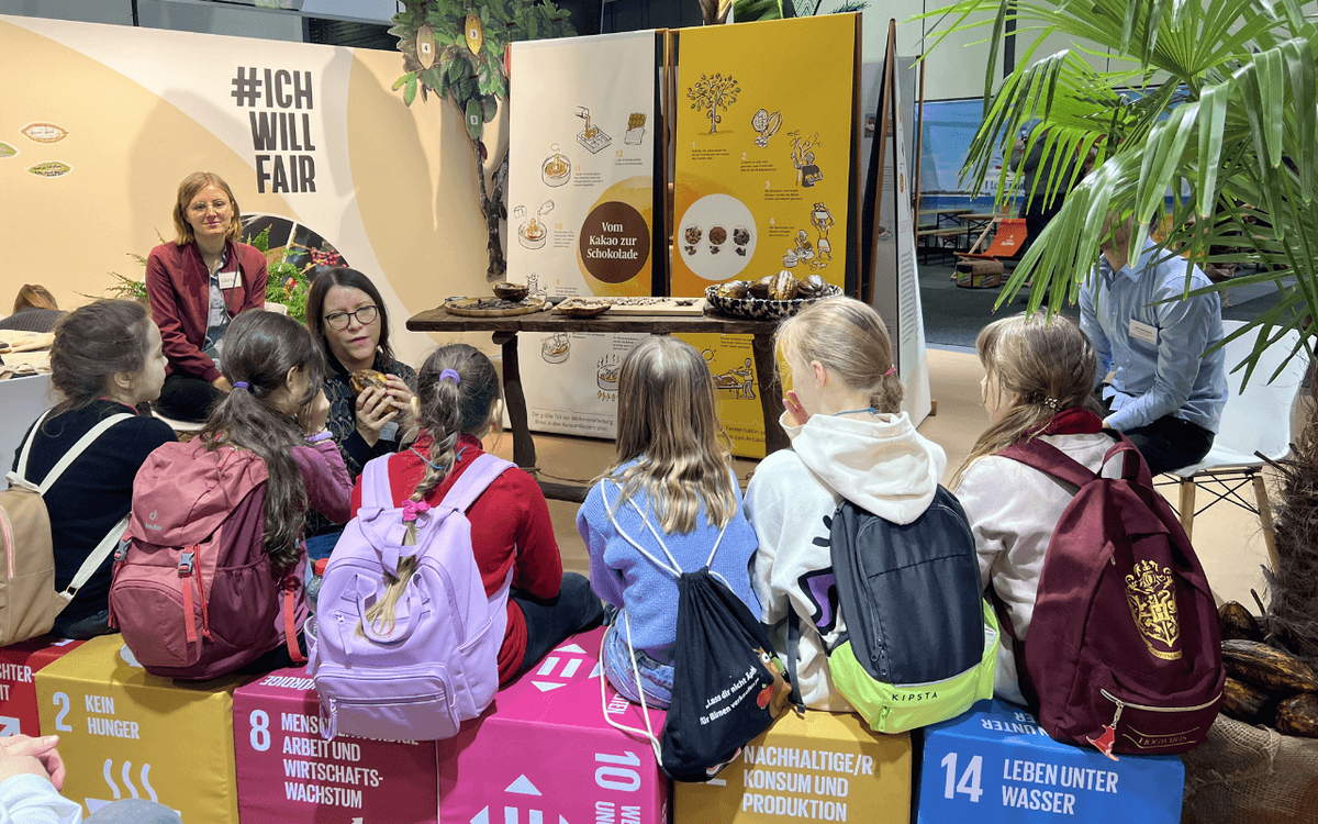 Kinder mit Rucksäcken sitzen vor einem Stand, der den Prozess von Kakao zu Schokolade erklärt, während eine Frau spricht.