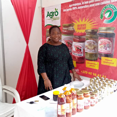 A woman stands smiling at a booth showcasing Agro Express products, including jars and bottles of sauces and pastes.