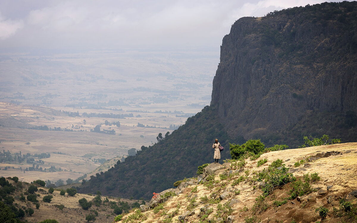 A person stands on a rocky hill, overlooking a vast, scenic landscape with a towering cliff and expansive fields below.