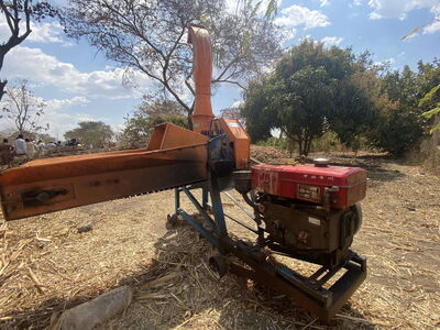 Eine große orangefarbene Maschine steht auf einem Feld unter Bäumen, während Menschen im Hintergrund arbeiten.