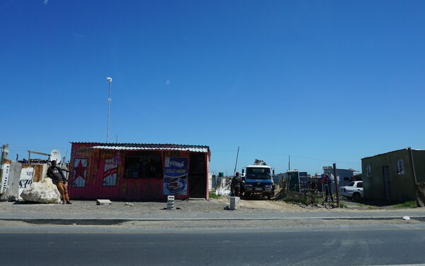 Ein kleines, bunt bemaltes Gebäude steht unter klarem Himmel neben einer Straße, während ein Mann davor lehnt.