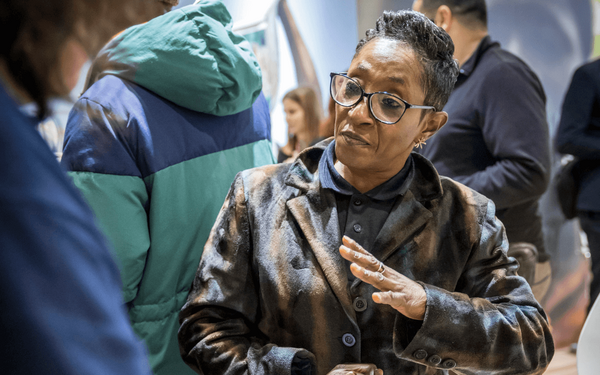 A woman in glasses and a patterned coat gestures while engaging in conversation at a social gathering.