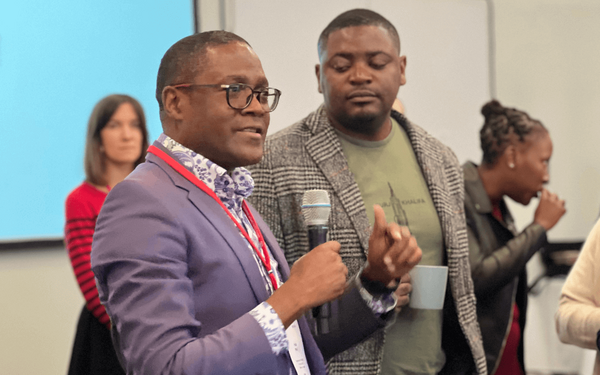 A man in a blue suit speaks into a microphone while others listen attentively in a conference setting.