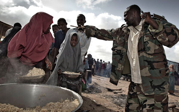 Ein Mann in Tarnkleidung überwacht die Essensverteilung an eine Gruppe von Menschen, darunter Kinder und Erwachsene.