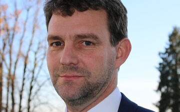 A man in a suit stands outdoors with trees in the background on a sunny day.
