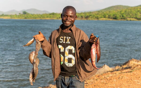 Ein lachender Mann steht am Seeufer und hält frisch gefangenen Fisch in beiden Händen hoch.