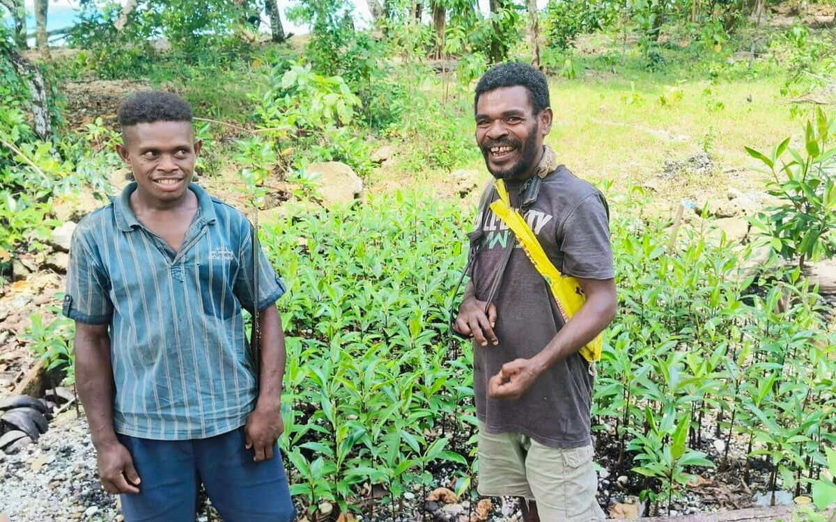 Two men stand smiling in a lush area dedicated to mangrove restoration, surrounded by young green plants.