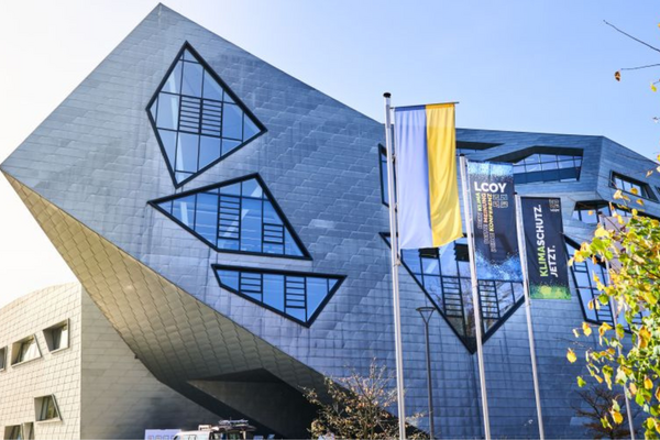 A modern, angular building with geometric windows stands beside flags at the LCOY event, surrounded by autumn trees.