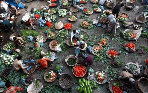 Vendors in a bustling Bangladeshi market sell vibrant vegetables displayed in woven baskets on the ground.