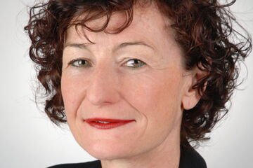 Eine Frau mit lockigem Haar und rotem Lippenstift blickt freundlich in die Kamera.