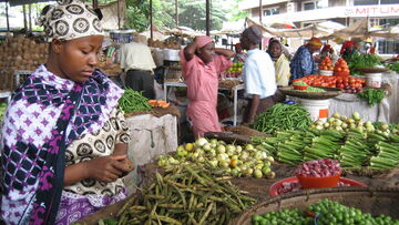 A woman in traditional attire inspects fresh vegetables at a bustling open-air market in Tanzania.