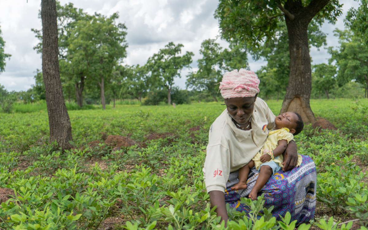 Eine Frau in Burkina Faso hält ein schlafendes Baby auf einem grünen Feld voller Pflanzen.