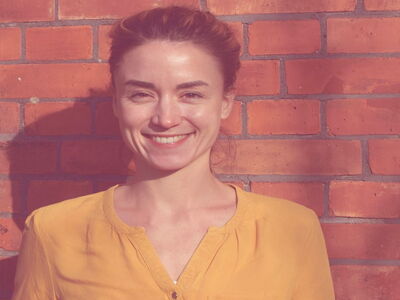 A woman in a yellow shirt smiles warmly while standing against a sunlit red brick wall.