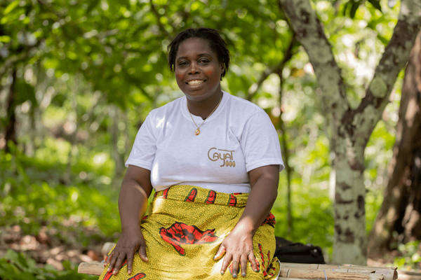 A woman in a white shirt and colorful skirt smiles while sitting outdoors, surrounded by lush greenery, promoting Fairtrade.