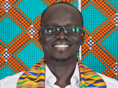 A smiling man wearing glasses and a colorful scarf stands against a vibrant patterned background.