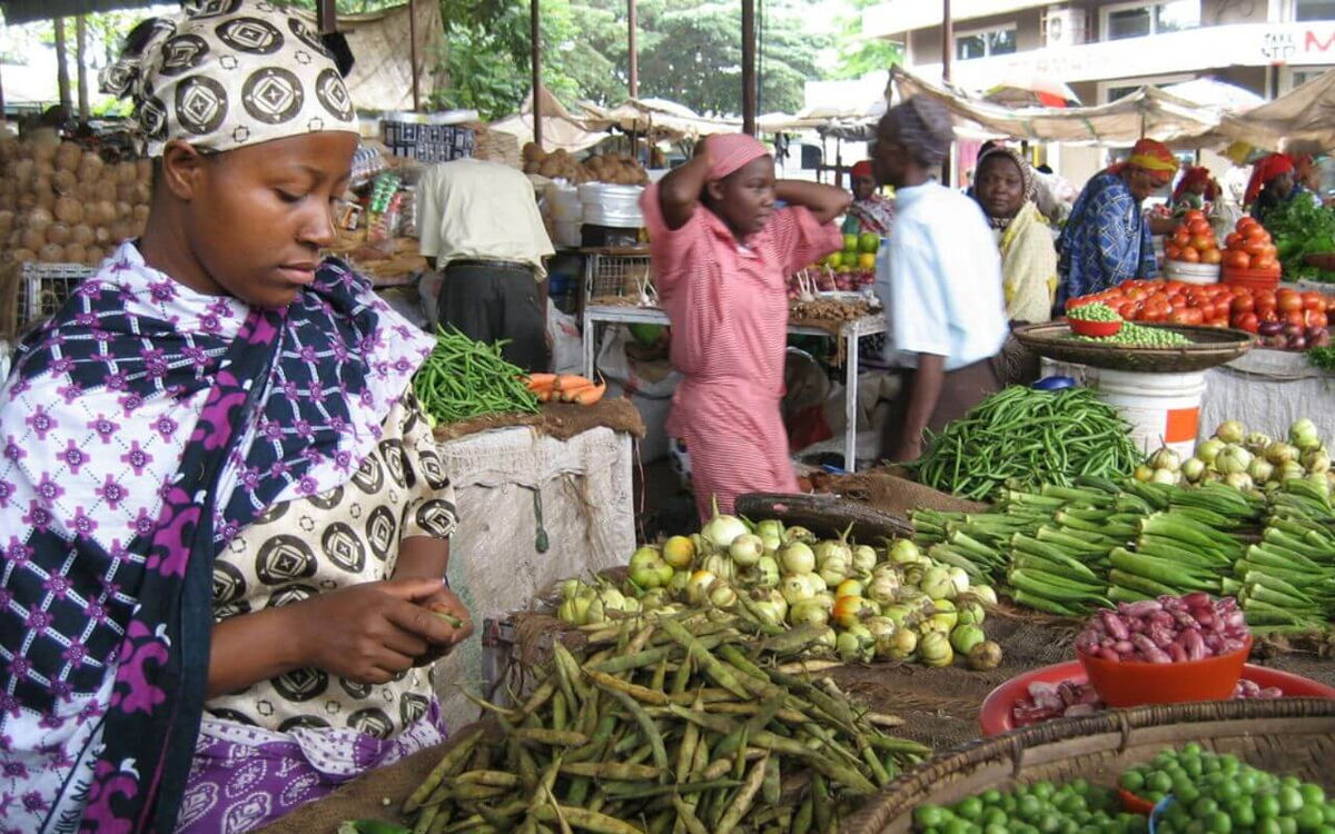 Eine Frau sortiert Gemüse auf einem belebten Marktstand, umgeben von bunten frischen Produkten und anderen Verkäufern.