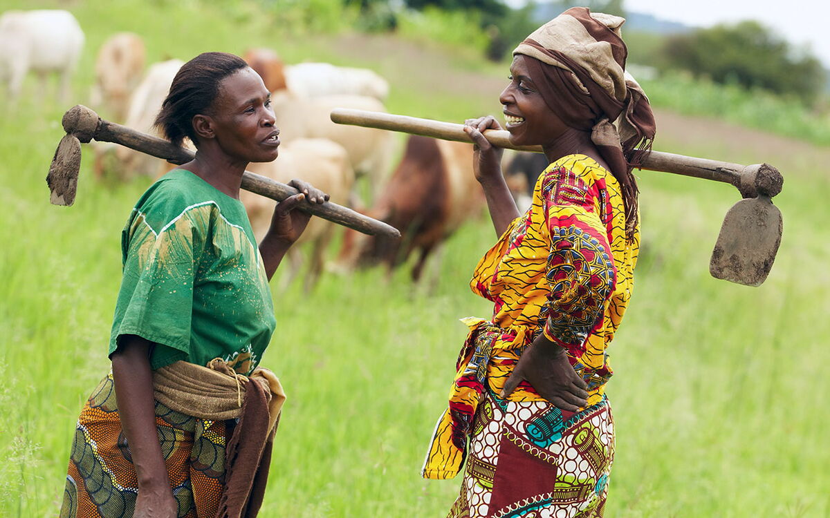 Two women in colorful attire chat happily while holding hoes over their shoulders in a lush green field.
