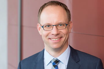 A smiling man in a suit stands against a red wall, exuding professionalism and confidence.