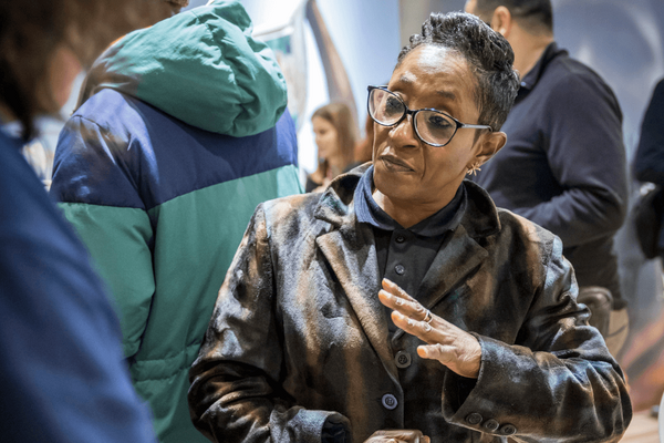 A woman in glasses and a patterned coat gestures while engaging in conversation at a social gathering.