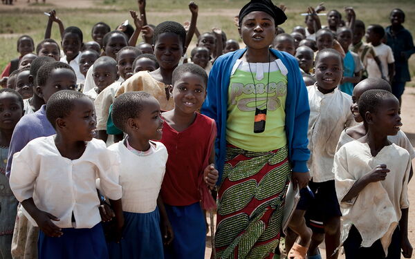 Eine Frau geht lächelnd mit einer Gruppe fröhlicher Kinder über einen Feldweg.