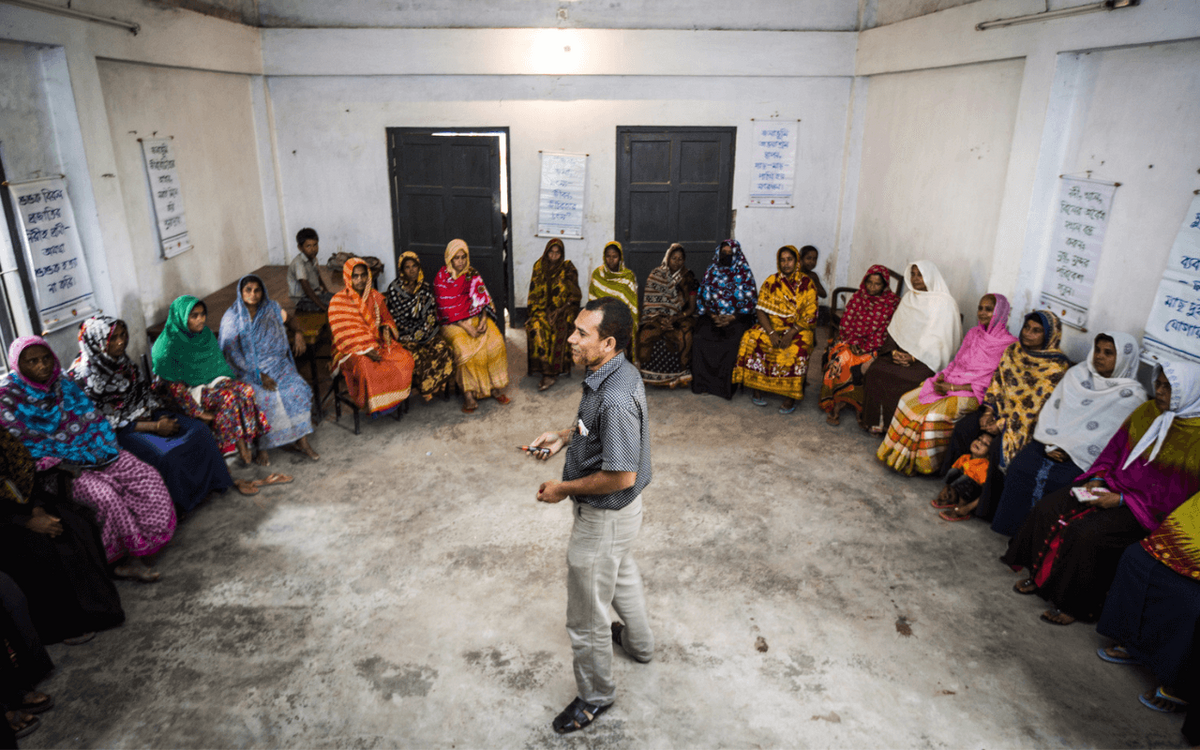 A man conducts a training session for a group of women seated in a circle in a Bangladesh community center.