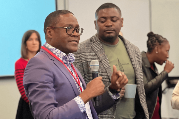 A man in a blue suit speaks into a microphone while others listen attentively in a conference setting.
