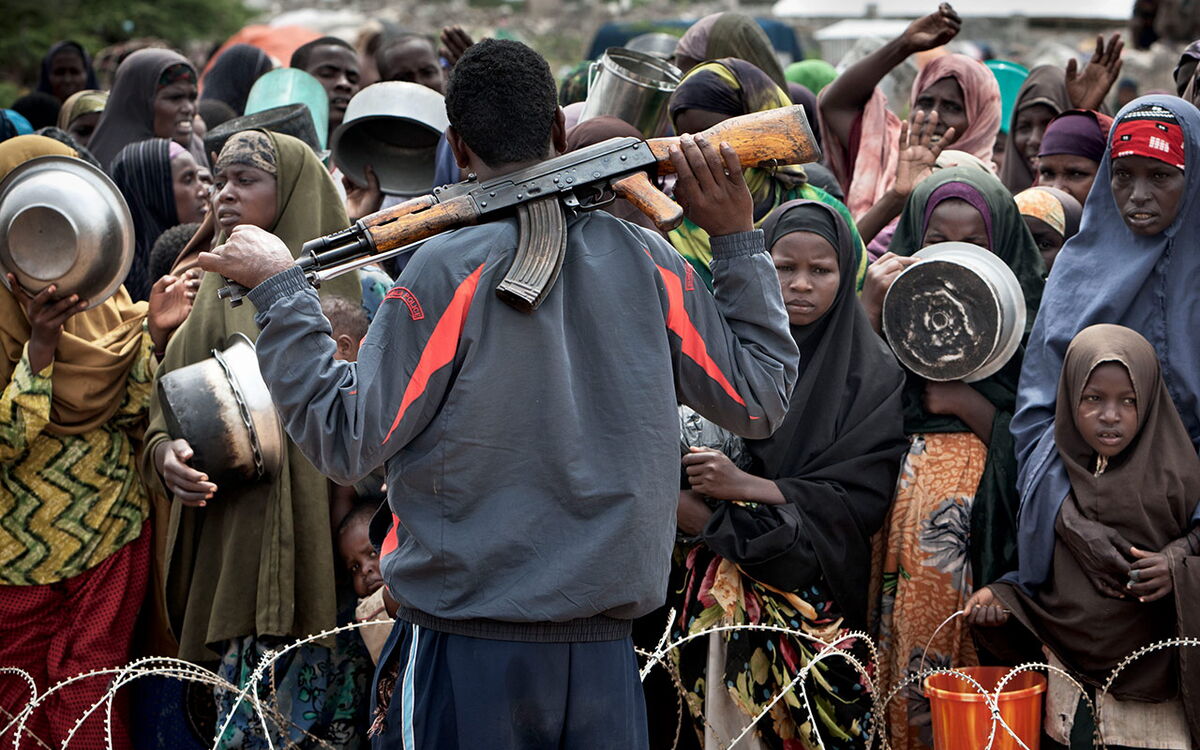 Ein bewaffneter Mann steht vor einer Menge von Menschen mit Kochtöpfen in der Hand hinter einem Stacheldraht.