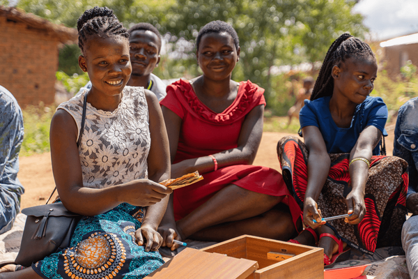 A group of smiling young people sit outdoors, engaging in a lively activity with a wooden box.