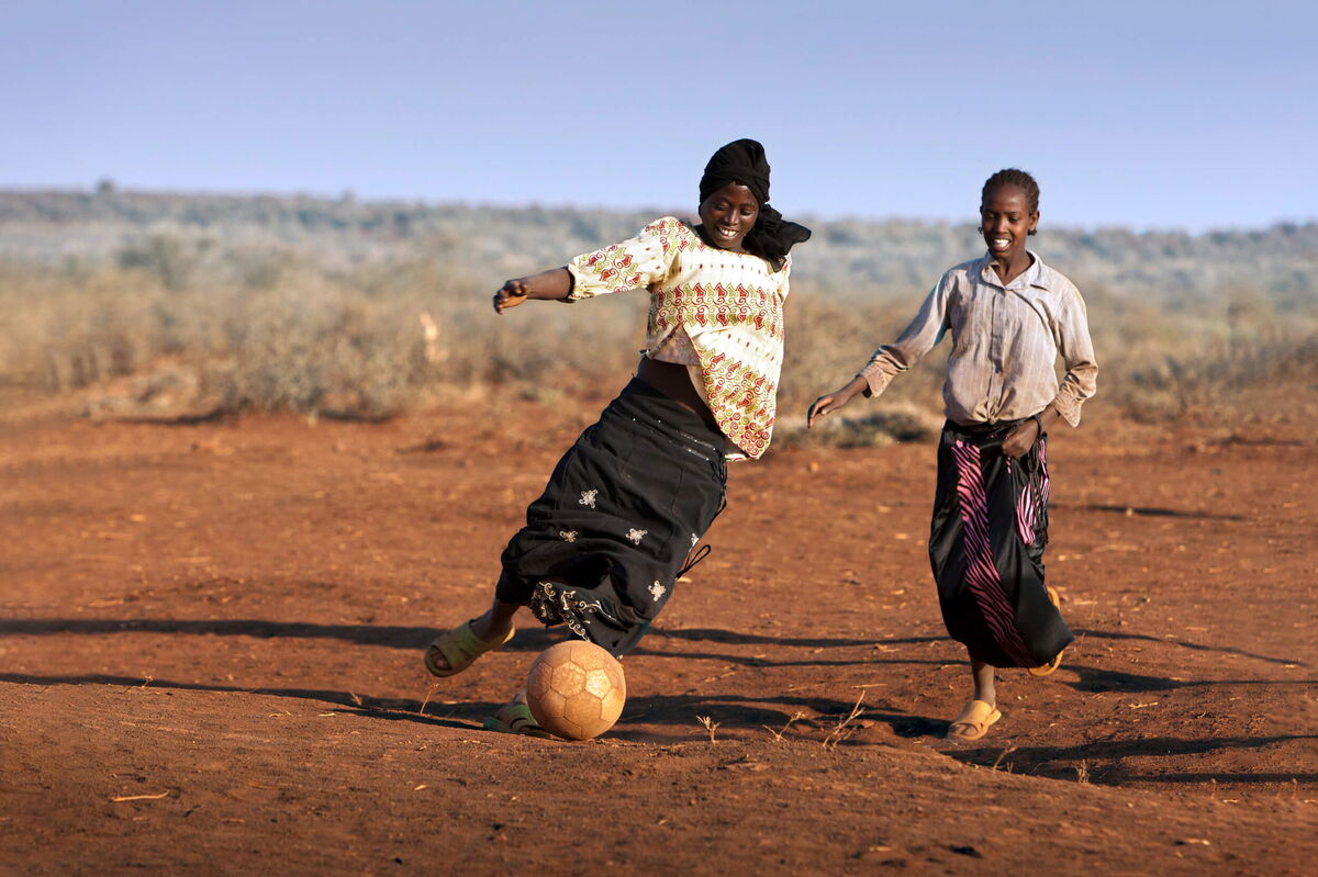 Two girls joyfully play soccer on a dusty field, capturing the spirit of playful perspectives.