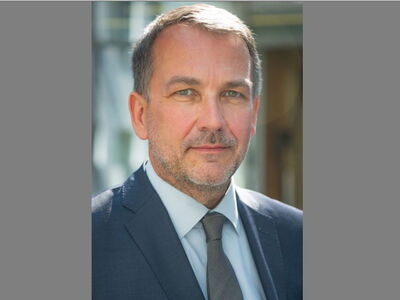 A man in a suit stands confidently outdoors with a blurred background.