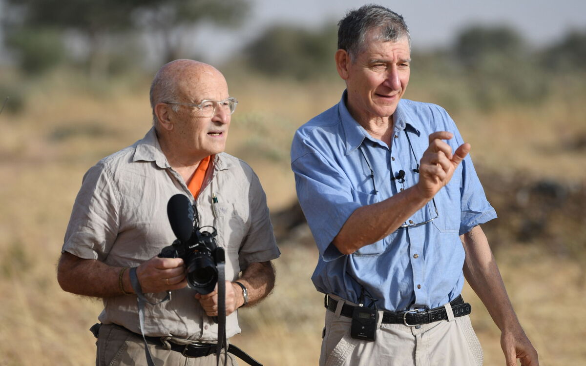 Zwei Männer stehen in der Natur Ghanas, einer hält eine Kamera, während der andere gestikuliert und spricht.