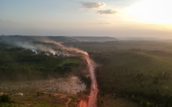 Ein staubiger Weg schlängelt sich durch eine grüne Landschaft, während Rauchfahnen in der Abenddämmerung aufsteigen.