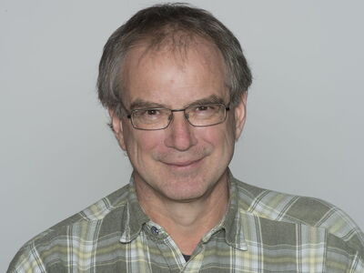 A man with glasses smiles warmly, wearing a plaid shirt against a neutral background.