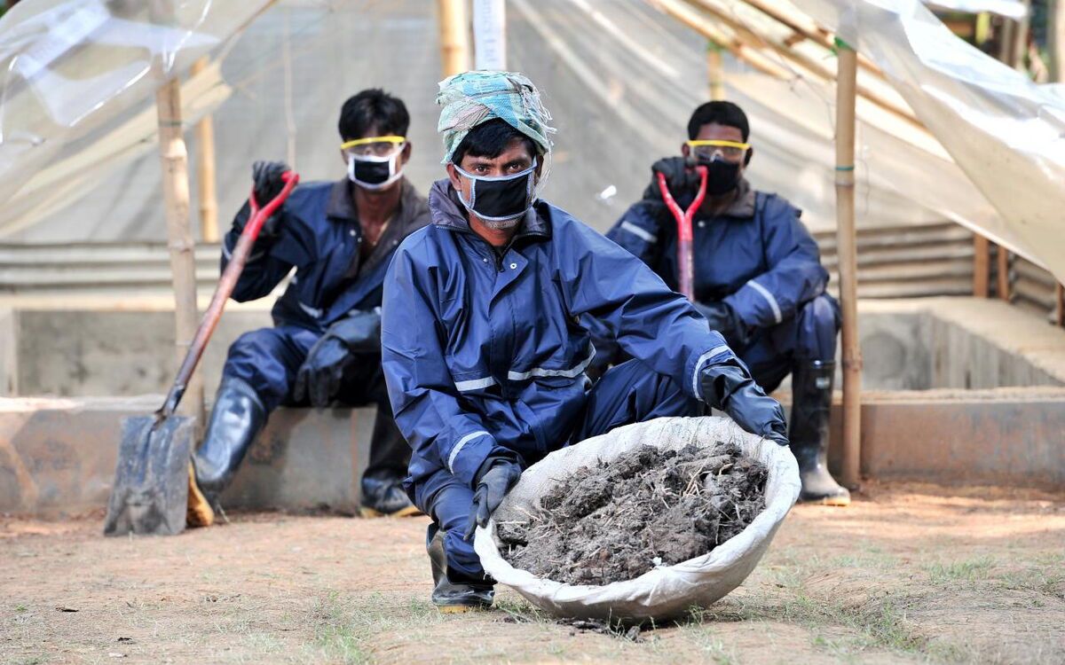 Drei Männer in Schutzanzügen arbeiten mit Schaufeln und Erde auf einer Baustelle im Freien.