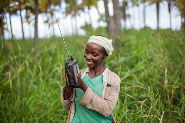 A woman in a green shirt smiles while holding a radio in a lush, green field in Tanzania.