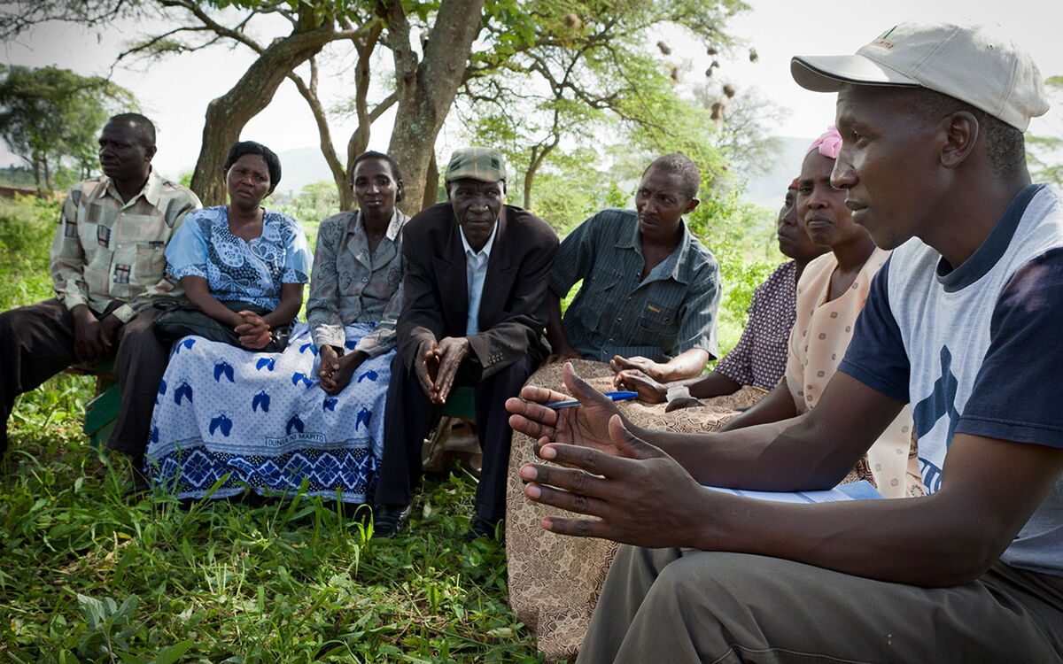 A group of people sit outdoors, attentively listening to a man speaking in a community meeting under a tree.