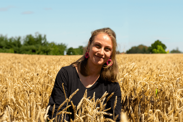 Eine Frau mit rosa Ohrringen lächelt in einem goldenen Weizenfeld unter blauem Himmel.