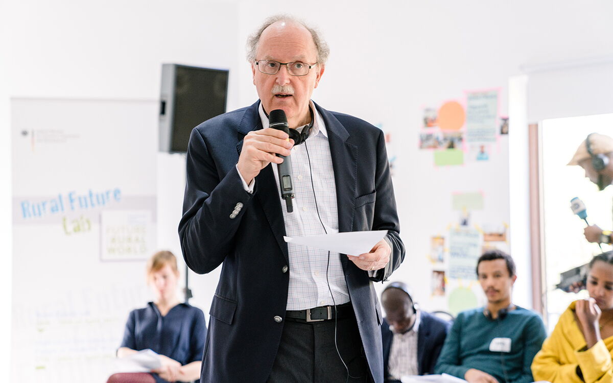 Ein Mann hält einen Vortrag und spricht in ein Mikrofon, während er ein Dokument vor einem Publikum präsentiert.