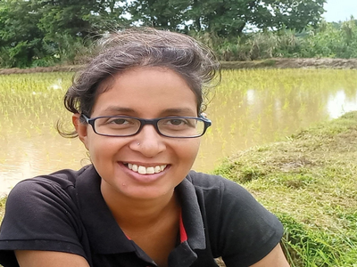 A woman in glasses smiles warmly while sitting outdoors near a lush, green field and water.