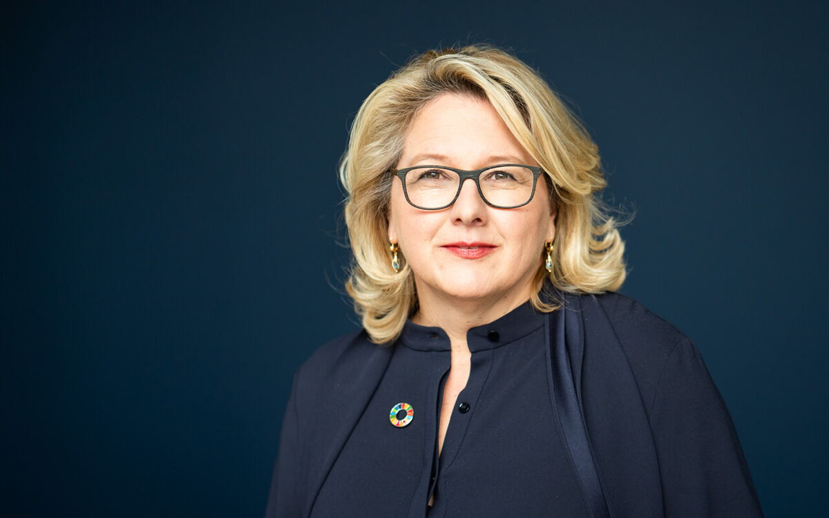 Eine Frau mit blonden Haaren und Brille lächelt vor einem dunklen Hintergrund, gekleidet in eine dunkle Bluse.